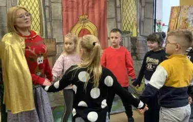 Na tle wnętrza pałacu kobieta przebrana za renifera w złotej pelerynie z dziećmi z publiczności tańczy w k&oacute;łeczku.