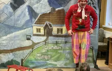 Na tle zimowej chaty w g&oacute;rach stoi aktor przebrany za renifera i ma otwarte mocno buzię z radości. Obok leżą czerowne sanki.