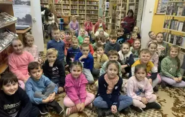 Dzieci dużą gromadą siedzą na dywanie, za nimi na krzesłach siedzą nauczyciele. 