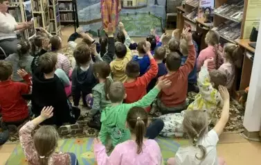 Dzieci siedzą na dywanie, niek&oacute;re z nich podnoszą rękę. Z przodu na tle zimowej chaty w lesie stoi aktor w czerwonej koszuli i ż&oacute;łtofioletowych spodniach i ma podniesione prawą rękę.