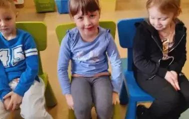 Troje dzieci siedzą na krzesełkach, na środku dziewczynka się delkiatnie uśmiecha.
