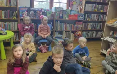 Dzieci siedzą na siedziskach i słuchają bajki, kt&oacute;rą czyta im bibliotekarka.