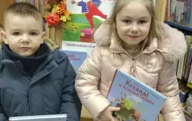 Brat z siostrą stoją obok siebie i trzymają nowe książeczki z wyprawki czytelniczej. Za nimi jest tablica z plakatem dotyczącym projektu
