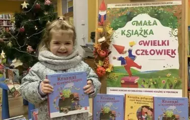 Uśmiechnięta dziewczynka  w blond włosach trzyma swoją nową książeczkę z wyprawki czytelniczej. Po prawej stronie stoi tablica korkowa z plakatem dotyczącym projektu.