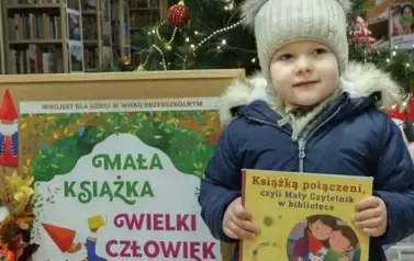  Dziewczynka w jasnej czapce trzyma swoją nową książkę z wyprawki. Za nią stoi korkowa tablica z plakatem dotyczącym projektu.