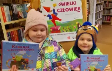 Brat z siostrą stoją obok siebie i trzymają nowe książeczki z wyprawki czytelniczej i karty Małego Czytelnika. Za nimi jest tablica z plakatem dotyczącym projektu