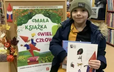 Chłopiec siedzi na krzesełku, trzyma nową książeczkę z wyprawki, zakładkę i kartę do wycięcia kostki do gry. Za nim stoi korkowa tablica z plakatem dotyczącym projektu.