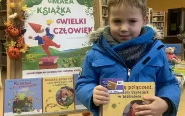 Chłopiec w jasnych włosach trzyma swoją nową książeczkę z wyprawki i kartę Małego Czytelnika.Za nim stoi tablica z plakatem projektu.