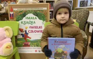 Dziewczynka stoi z nową książeczką z wyprawki. Obok jest tablica korkowa z plakatem dotyczącym projektu.