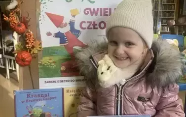 Uśmiechnięta dziewczynka w jasnej czapce trzyma swoją nową książkę z wyprawki. Za nią stoi korkowa tablica z plakatem dotyczącym projektu.