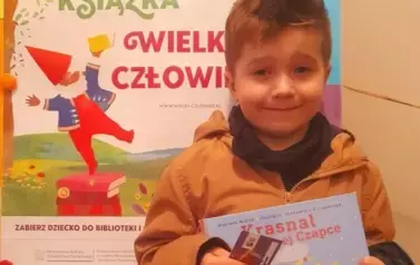 Chłopiec w jasnych włosach trzyma swoją nową książeczkę z wyprawki i kartę Małego Czytelnika.Za nim stoi tablica z plakatem projektu.
