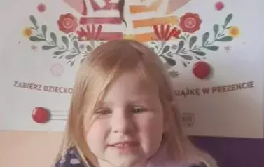 Dziewczynka w blond włosach trzyma swoją nową książeczkę z wyprawki i kartę Małego Czytelnika.Za nią stoi tablica z plakatem projektu. 