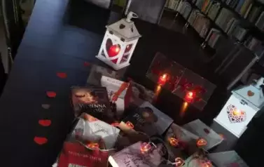 Na stole są wyłożone książki o tematyce miłosnej przykryte lampkami i otoczone serduszkami. Za nimi stoją zapalone dwa lampiony i dwie świeczki.