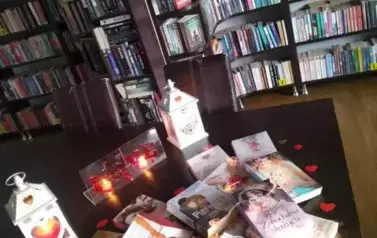 Na stole są wyłożone książki o tematyce miłosnej przykryte lampkami i otoczone serduszkami. Za nimi stoją zapalone dwa lampiony i dwie świeczki.