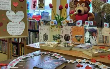Na biurku są wyłożone książki o tematyce miłosnej, na stole obok jest na środku książka otoczona lizakami w serduszka i płatkami r&oacute;ż. Nad wisi napis "Walentynki". Z lewej strony stoi korkowa tablica z grafiką walentynkową