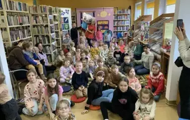 Bardzo liczna grupa dzieci, opiekun&oacute;w i rodzic&oacute;w siedzi na dywanie, a z tyłu jest aktorka Bajanka, kt&oacute;ra trzyma maskotkę Kicię Kocię