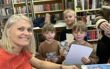 Dwie dziewczynki i chłopiec stoją przy stoliku pisarki aby zakupić książeczki. Trzymają w rączkach kartkę i kicie kocie z papieru. Pisarka się ładnie uśmiecha.