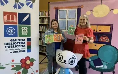 Na tle bajkowego pokoju przy Kici Koci stoi Bajanka i kierowniczka biblioteki. Obie trzymają po dwie książeczki bajkowe. Z lewej strony stoi logo biblioteki.