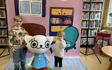 Na tle bajkowego pokoju przy Kici Koci stoi dziewczynka i chłopiec.