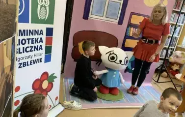 Na tle bajkowego pokoju przy Kici Koci siedzi chlopiec w ciemnych włosach trzymając ją za rękę. Obok stoi Bajanka i patrzy.