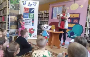 Na tle bajkowego pokoju obok Kici Koci stoi przy stoliku Bajanka w okularach i rękawiczkach robiąc eksperyment. Po lewej stronie stoi dziewczynka i patrzy. 