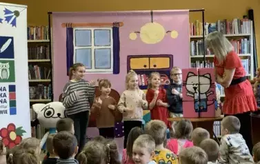 Na tle bajkowego pokoju przy Kici Koci stoi pięcioro dzieci i patrzą na Bajankę, kt&oacute;ra im coś pokazuje rękami. Obok na stoliku stoją klocki Kici Koci.