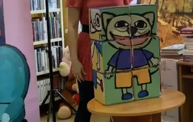 Bohaterka bajki Bajanka stoi wyprostowana i uśmiechnięta przy stoliku na kt&oacute;rym są duże klocki Kici Koci. 