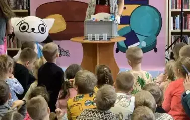 Na tle bajkowego pokoju stoi przy stoliku do eksperyment&oacute;w Bajanka i pokazuje ręką w białej rękawiczce w kierunku Kici Koci. Przed tą scenką siedzą dzieci.