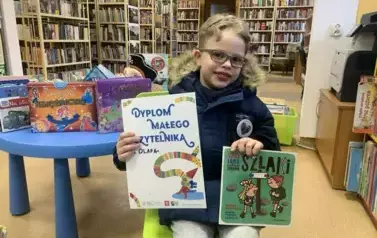 Chłopiec w blond włosach i okularkach siedzi na krzesłku trzymając dyplom i wypożyczoną książeczkę.