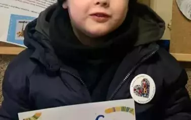 Chlopiec w czarnej czapce stoi przy korkowej tablicy z grafikami dotyczącymi akcji MAŁA KSIĄŻKA WIELKI CZŁOWIEK. Trzyma w rękach dyplom.