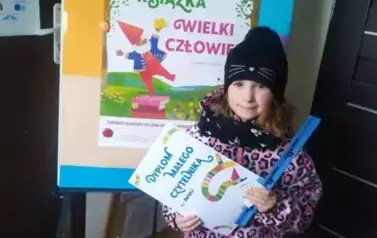 Dziewczynka w czarnej czapce stoi przy tablicy z plakatem MAŁA KSIĄŻKA WIELKI CZŁOWIEK. Trzyma w rękach dyplom i opaskę na rękę.