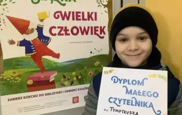 Chłopiec stoi przy drzwiach na kt&oacute;rych wisi plakat MAŁA KSIĄŻKA WIELKI CZŁOWIEK. Trzyma w rękach dyplom i się uśmiecha.