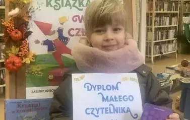 Dziewczynka w blond włosach stoi przy tablicy z plakatem dotyczącym akcji MAŁA KSIĄŻKA WIELKI CZŁOWIEK. Trzyma w rękach dyplom i kartę Małego Czytelnika.
