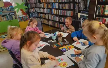 Dzieci przy stole obklejają plasteliną na kartkach kolorowe koguciki.