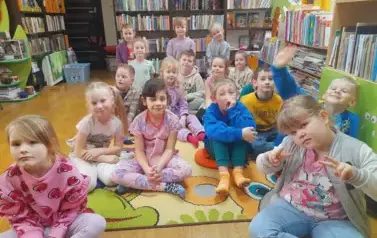 Dzieci siedzą na dywanie i słuchają bajki, kt&oacute;rą czyta im bibliotekarka.