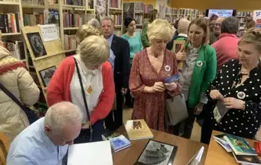 Przy biurku Janusz Szalewski podpisuje zakupione książki Czytelnikom, kt&oacute;rzy stoją w kolejce.