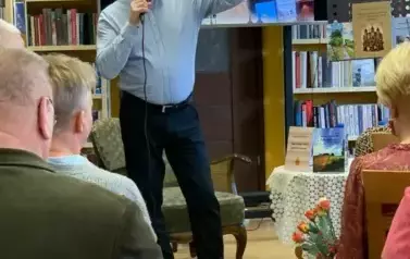 Janusz Szalewski ma podniesione rękę w kt&oacute;rej pokazuje swoją książkę . Za nim jest duży ekran z wyświetlonym plakatem dot. spotkania. Czytelnicy patrzą z zainteresowaniem.