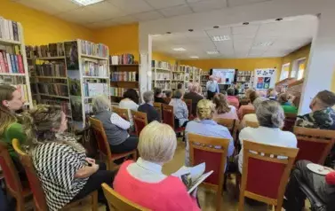 Biblioteka pełna publiczności siedzącej na krzesłach, z przodu autor opowiada o swojej tw&oacute;rczości.