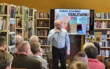 Czytelnicy patrzą w stronę autora, kt&oacute;ry opowiada o swojej tw&oacute;rczości. Za Nim wisi duży ekran z wyświetlonym plakatem dotyczącym spotkania.