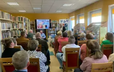 Biblioteka pełna publiczności siedzącej na krzesłach.  z przodu autor siedzi na fotelu obok stoi Stanisław Sierko prowadząc  dyskusję. Na środku nad wszystkimi wisi duży ekran z wyświetlonym plakatem dotyczącym spotkania.