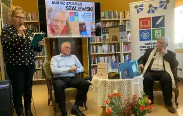 Przy okrągłym stoliku na kt&oacute;rym są wystawione książki autora siedzą Janusz Szalewski i Stanisław Sierko, kt&oacute;ry prowadził to spotkanie. Po lewej stronie czyta do mikrofonu wiersz żona pisarza. U g&oacute;ry wisi duży ekran z wyświtlonym plakatem dotyczącym spotka
