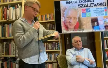 Autor siedzi na fotelu, nad Nim wisi duży ekran z wyświetlonym plakatem dotyczącym spotkania. Z lewej strony stoi kobieta z mikrofonem czytając wiersz Autora.