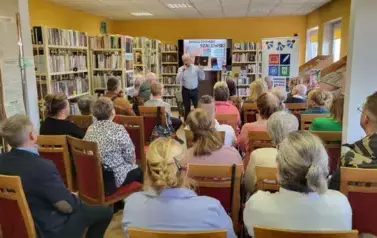 Z przodu autor opowiada pokazując swoją książkę, za Nim wisi duży ekran z wyświetlonym plakatem dotyczącym spotkania. Cała biblioteka pełna Cztelnik&oacute;w siedzących na krzesłach.