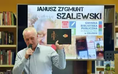 Janusz Szalewski ma podniesione rękę w kt&oacute;rej pokazuje swoją książkę pt &quot; Wielka Księga Szalewskich&quot;. Za nim jest duży ekran z wyświetlonym plakatem dot. spotkania.