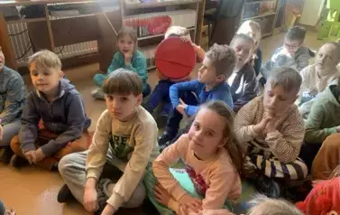 Dzieci siedzą na dywanie i siedziskach i patrzą na bibliotekarkę, kt&oacute;ra im opowiada o książce i o baśniach Andresena.
