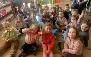 Dzieci siedzą na dywanie i słuchają bajki jaką czyta im bibliotekarka. Niekt&oacute;re z nich podnosza rączkę w celu chęci zabrania głosu.