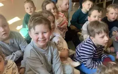 Dzieci siedzą na dywanie i słuchają bajki, kt&oacute;rą czyta im bibliotekarka, jeden chłopiec się szeroko uśmiecha. Z tyłu na krzesłach siedzą ich Panie.