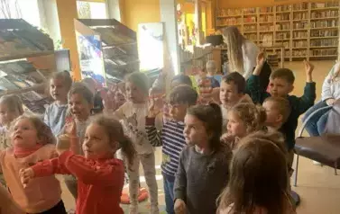 Dzieci stoją w rozsypce i niekt&oacute;re z nich mają wyciągnięte ręce do g&oacute;ry naśladując bibliotekarkę, kt&oacute;ra pokazuje im ćwiczenie.