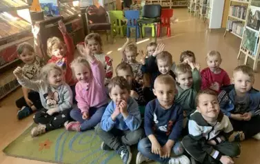 Dzieci siedzą na dywanie i słuchają bajki jaką czyta im bibliotekarka. Niekt&oacute;re z nich podnoszą rączkę w celu chęci zabrania głosu.