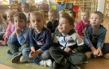 Dzieci siedzą na dywanie i z zaciekawieniem patrzą na bibliotekarkę, kt&oacute;ra czyta im bajkę.